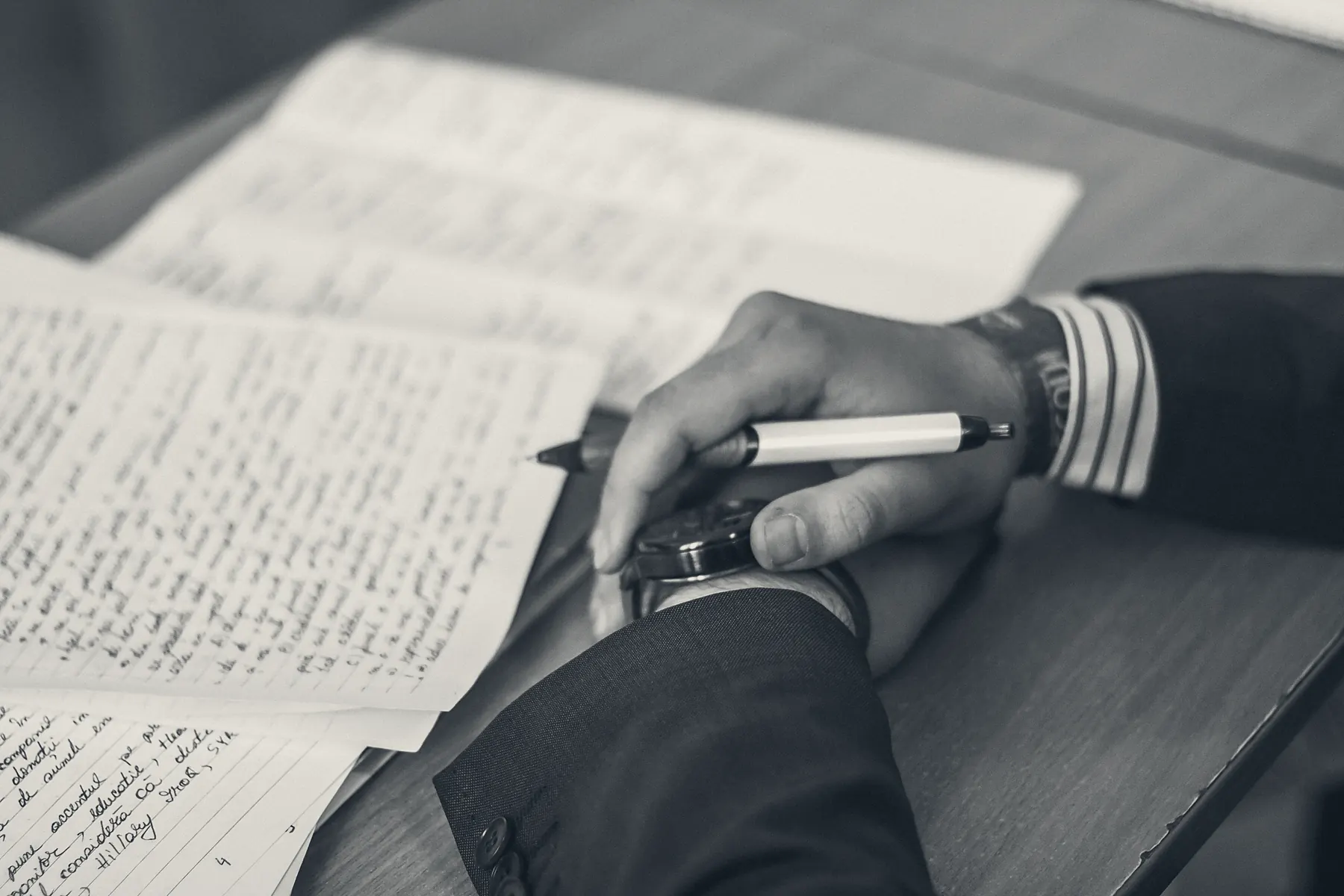 A hand resting a pen on a page of handwritten working notes, photographed close and in monochrome.